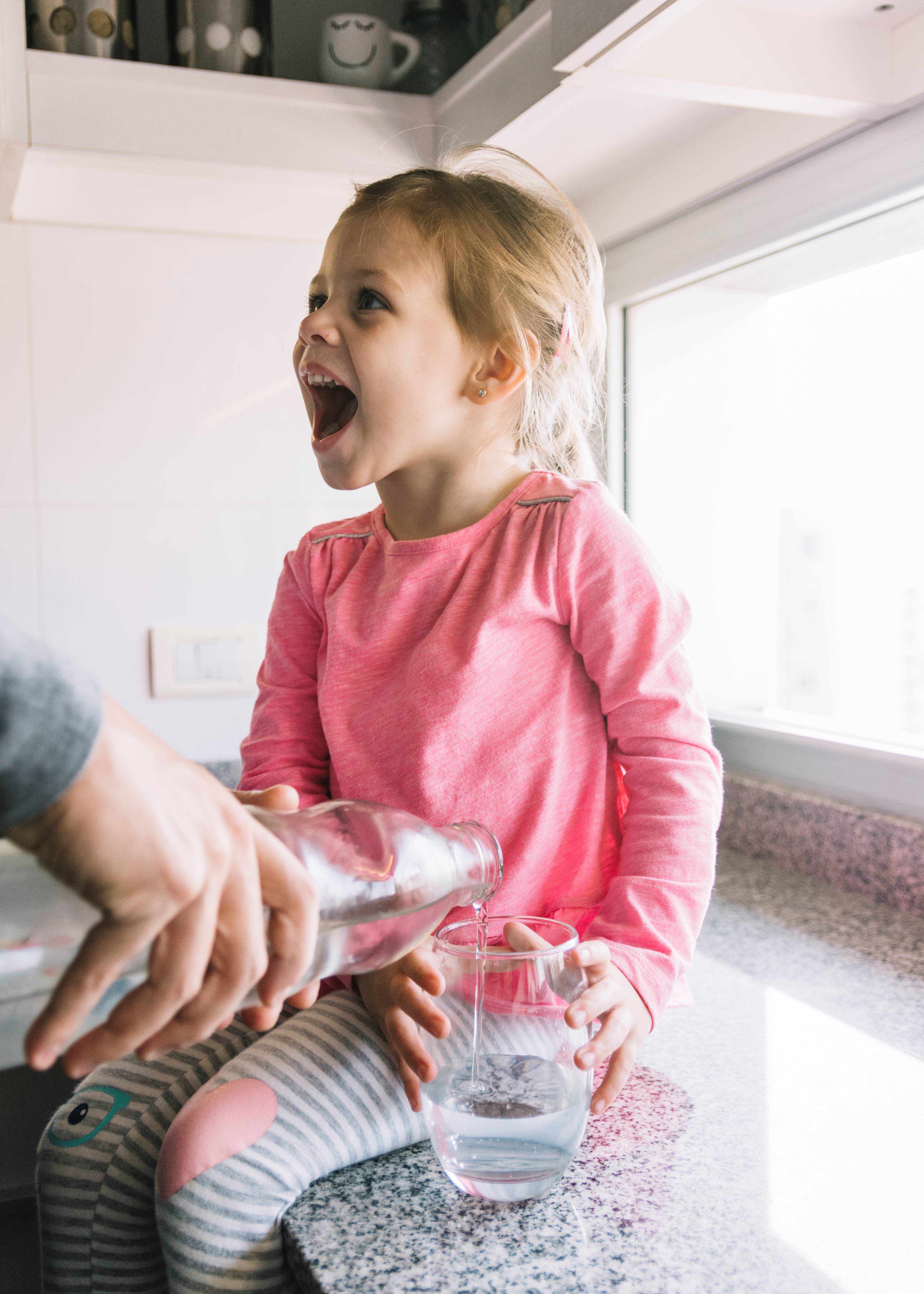 person-s-hand-pouring-water-glass-while-girl-sitting-kitchen-counter2